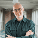 Bald man with glasses and a beard, wearing a dark green button-up shirt, stands smiling with arms crossed in a brightly-lit corridor.