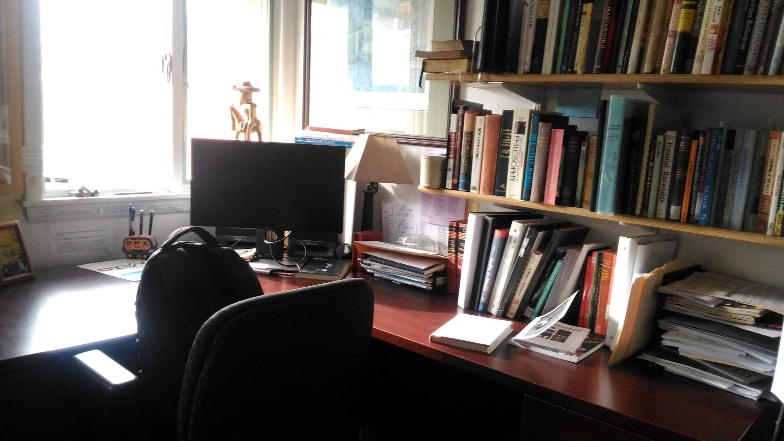 A sunlit home office with a computer, lamp, and bookshelves filled with books. Papers and folders are organized on the desk, and a backpack hangs on the chair. A window lets in natural light.