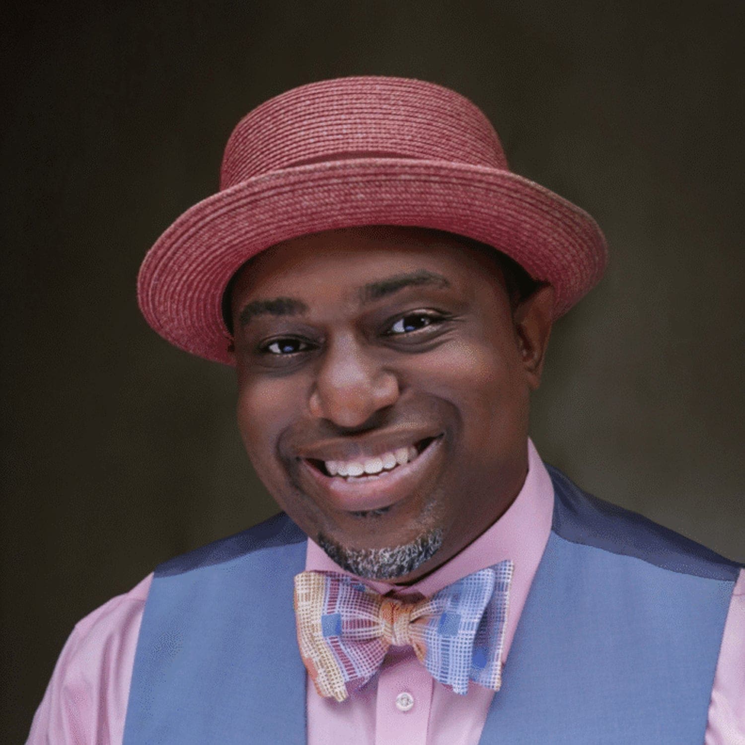 A smiling man wears a red hat, pink shirt, blue vest, and colorful bow tie, posed against a dark background.