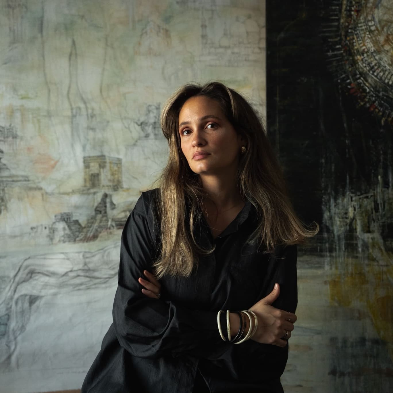 A woman with long hair wearing a black shirt stands with arms crossed in front of abstract art, looking slightly to the side.