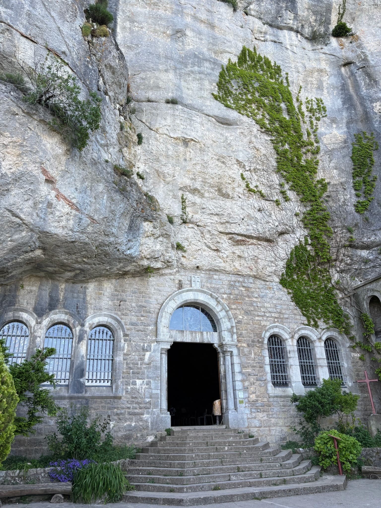 Stone church with arched windows built into a rocky cliff, greenery growing on the rock face, and steps leading up to the entrance.