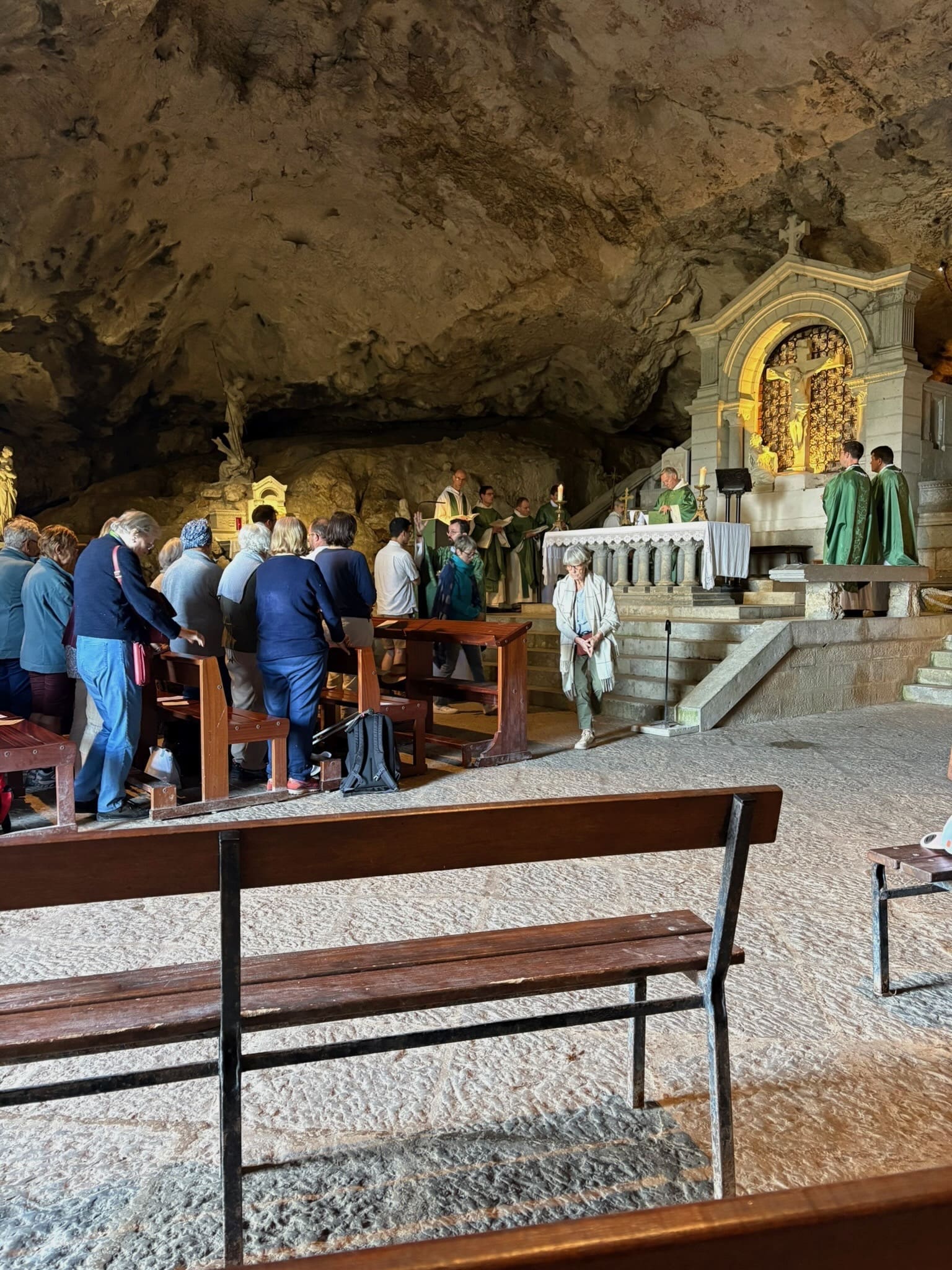 A group of people attend a religious service inside a rocky cave. Several clergy in green robes stand at an altar, while attendees pray or sit on benches facing them. A golden-lit shrine is visible in the background.