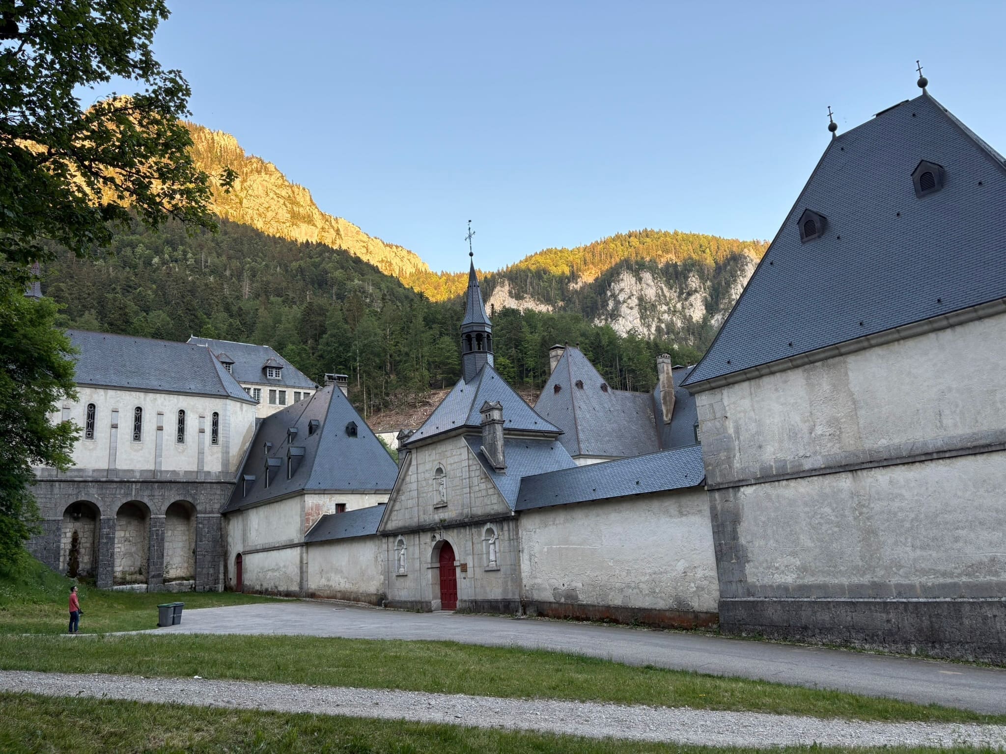 Stone buildings with gray roofs sit at the base of a tree-covered mountain. A person stands near a walkway, and warm sunlight illuminates the mountain in the background.
