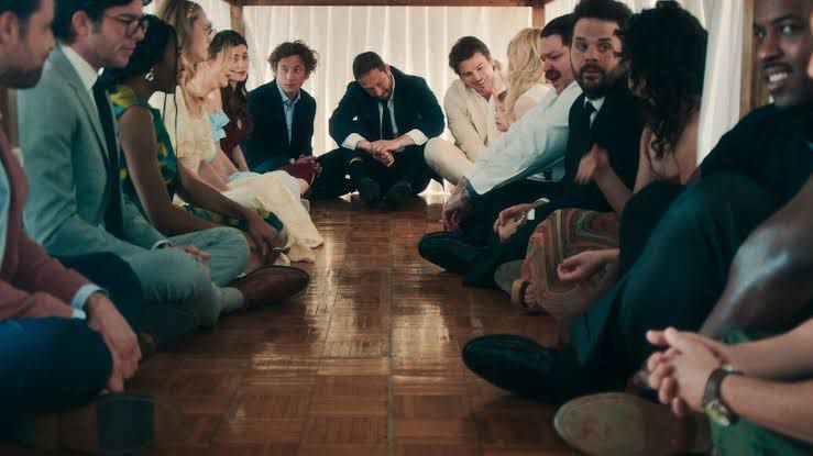 A group of people dressed formally sit facing each other in two rows on a wooden floor, appearing serious and engaged, with light streaming in from behind.