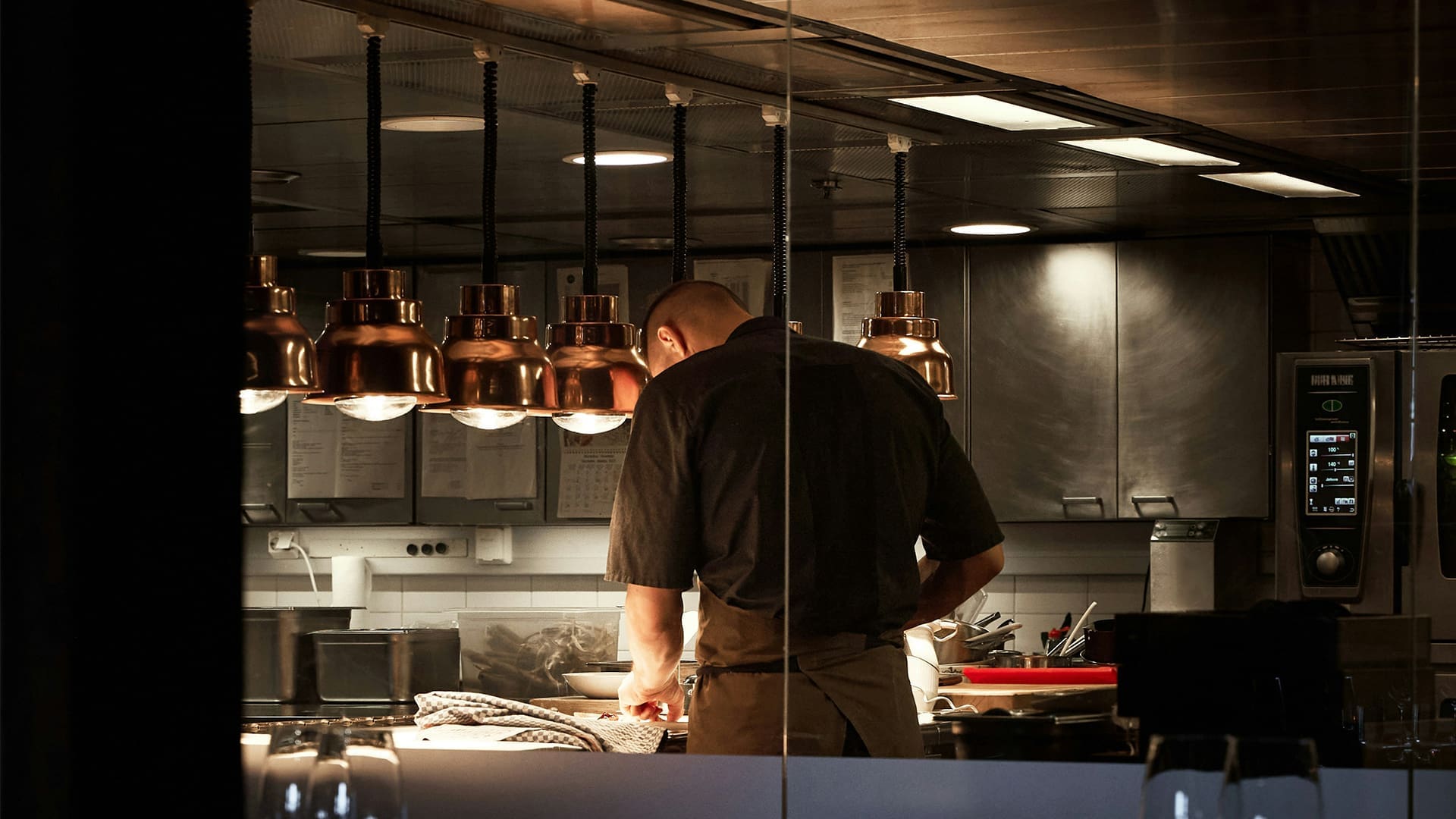A chef stands under warm hanging lights, preparing food in a professional stainless steel kitchen with various equipment, utensils, and notes visible in the background.