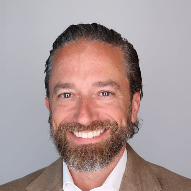 Smiling man with brown hair and beard, wearing a tan blazer and white shirt, posed in front of a plain gray background.