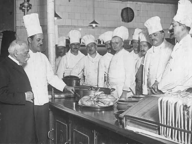 A group of chefs in white uniforms and hats stand around a kitchen counter with food, while two men in dark suits talk to them. There are pots, pans, and roasted poultry on the counter.