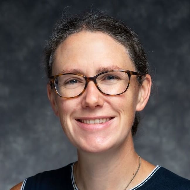 A person with light skin, glasses, and brown hair pulled back is smiling at the camera. The background is dark and blurred.