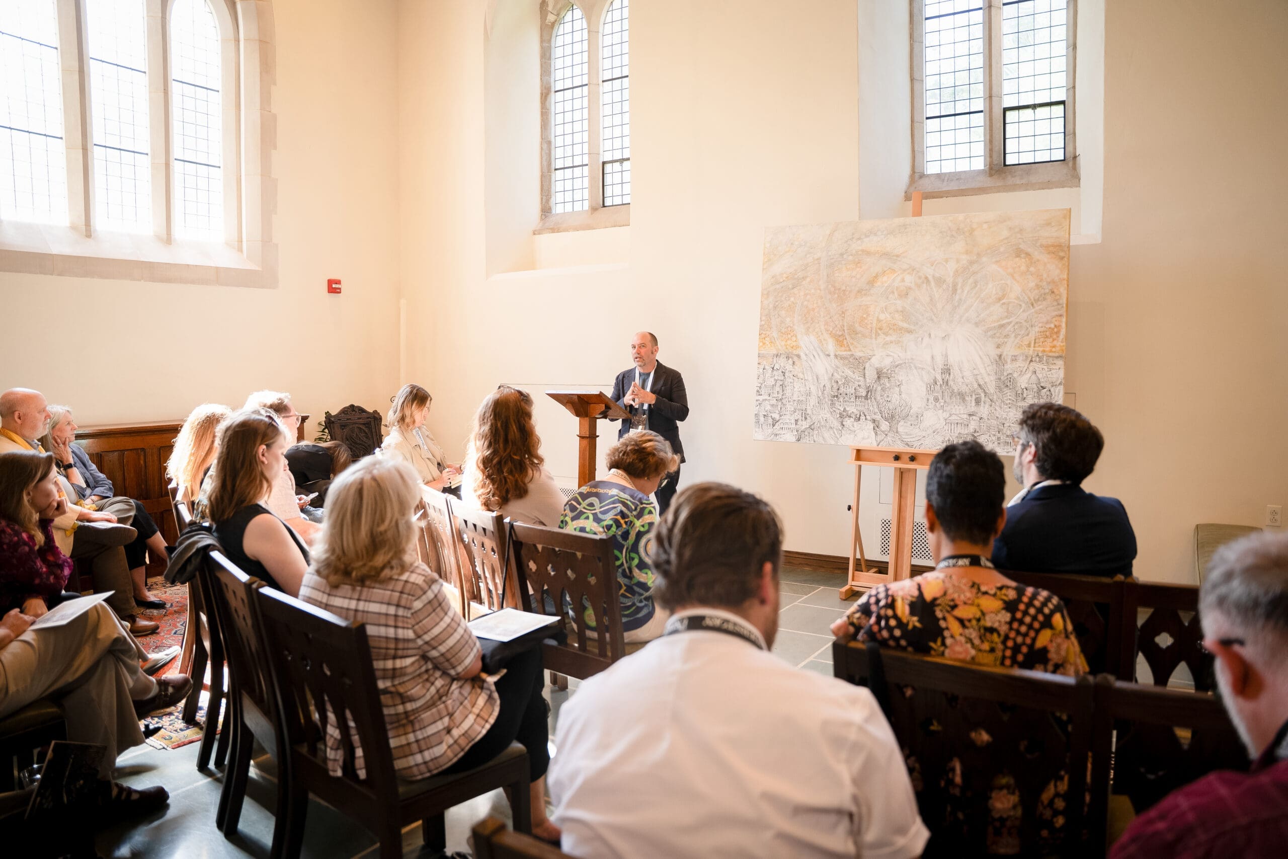 A man speaks at a podium in a sunlit room to a seated audience. Large windows let in natural light, and a large artwork on an easel stands beside the podium.