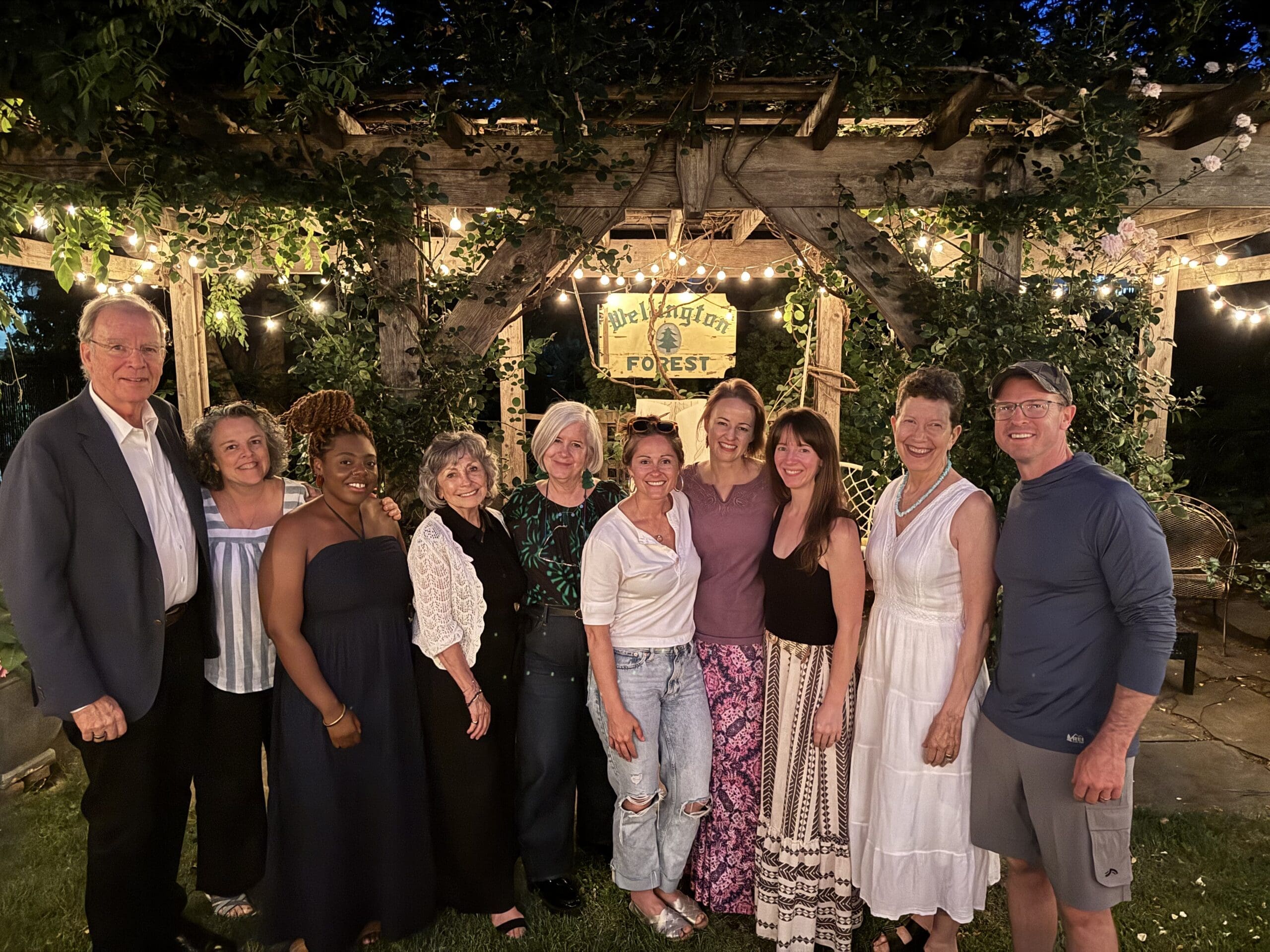 Ten people stand smiling together outdoors at night under string lights and greenery, with a wooden pergola and a sign reading The Magic Forest in the background.