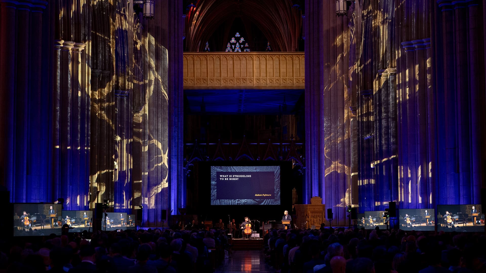 A large, ornate hall with tall arches is illuminated by golden lights. Multiple screens show a speaker at a podium. The center screen reads WHAT IF INTELLIGENCE IS A RED HERRING?.