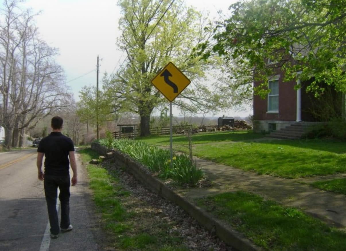 A person walks along a quiet road near a yellow curvy road sign, with a brick house and green trees on a sunny day.