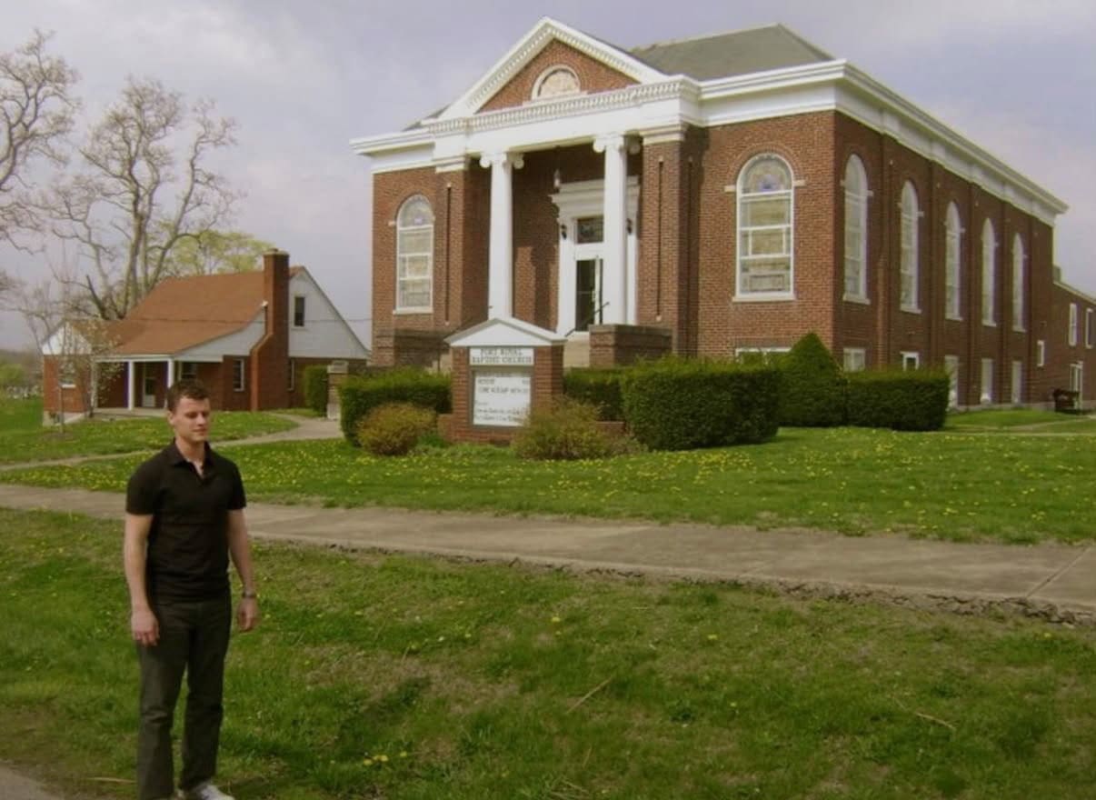 A man in a black shirt stands on a sidewalk in front of a large red-brick church with white columns and arched windows, surrounded by green grass and trees.