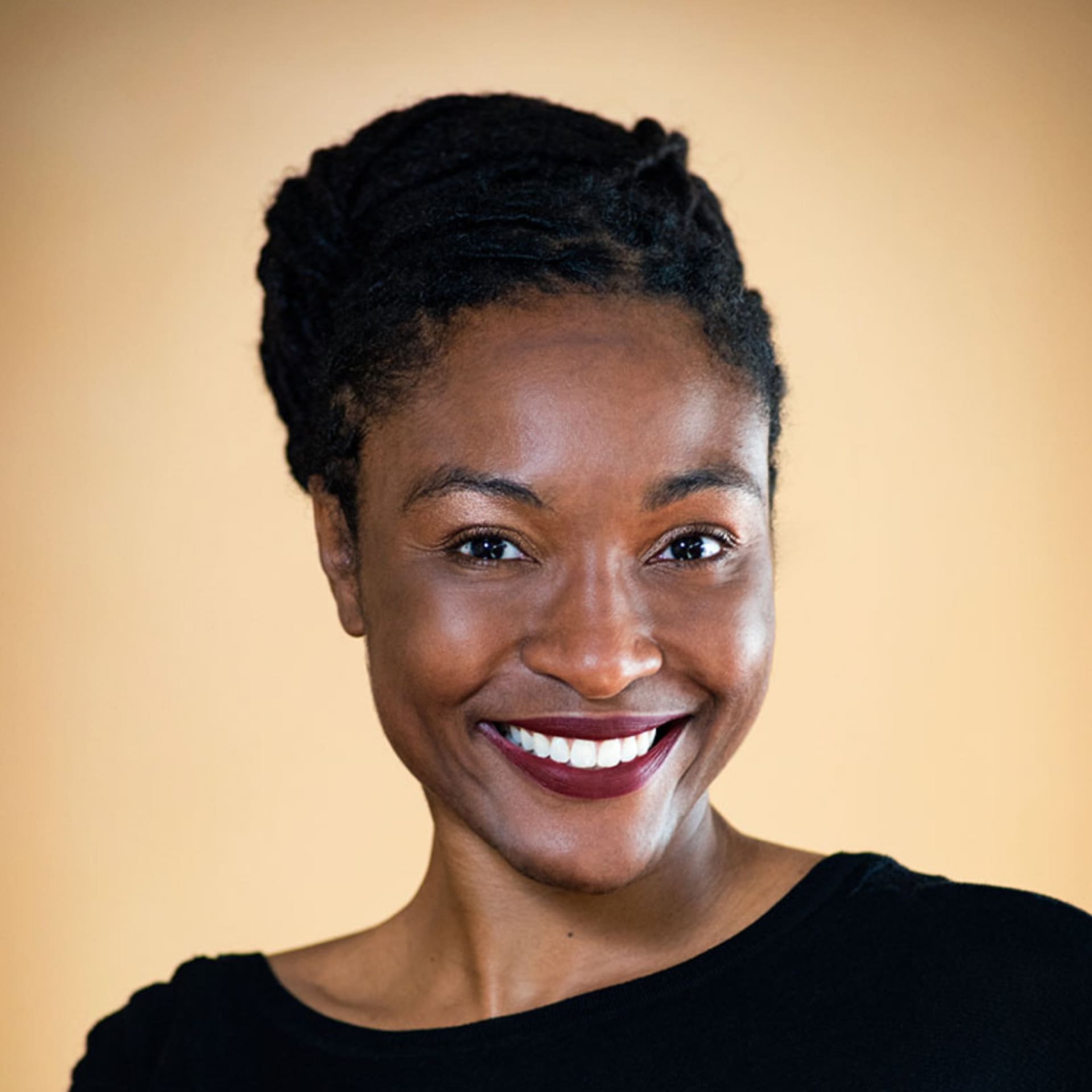 A smiling woman with dark braided hair pulled back, wearing a black top, stands in front of a soft beige background.