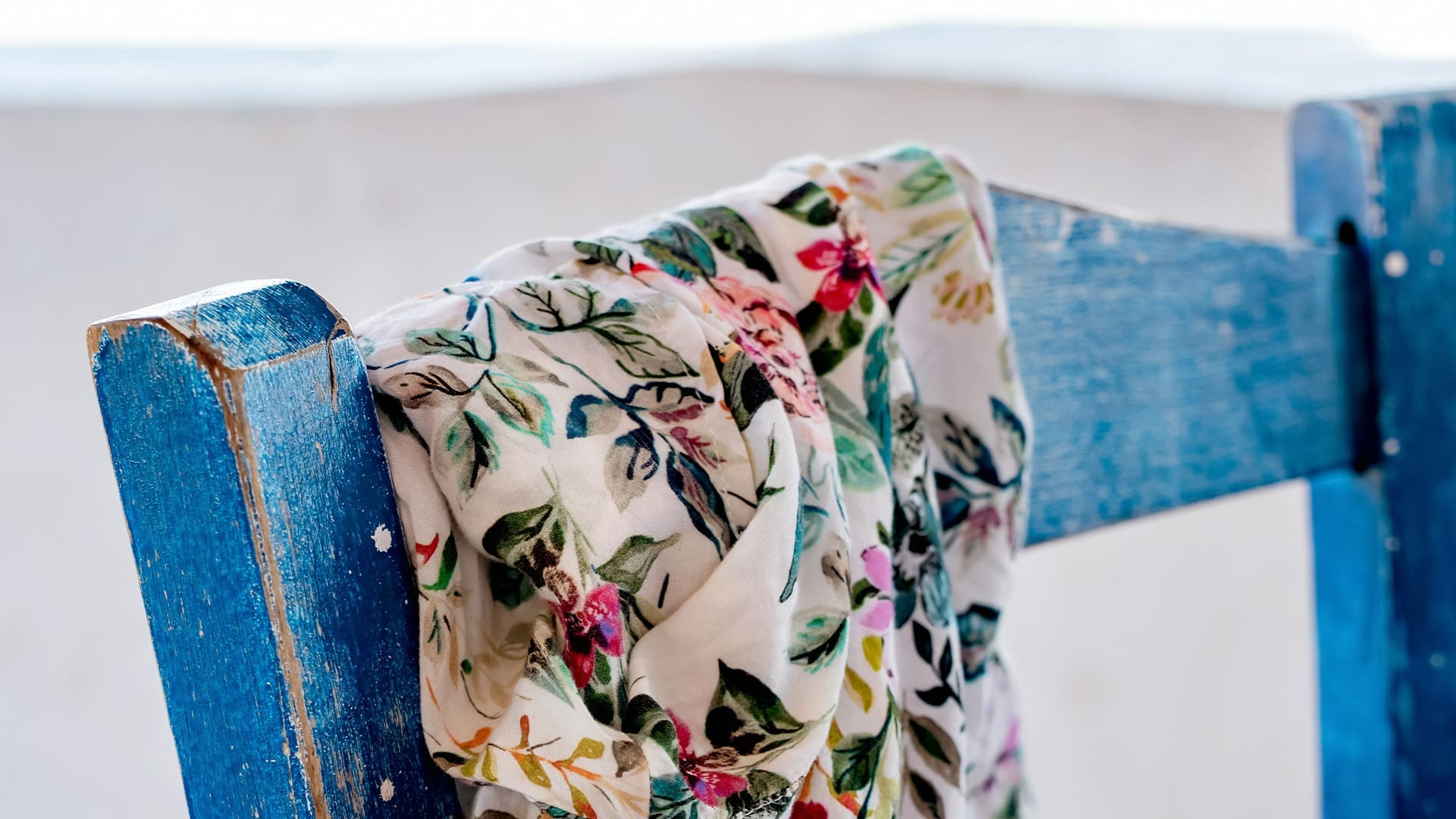 A colorful floral scarf draped over the back of a weathered blue wooden chair, with a blurred light background.