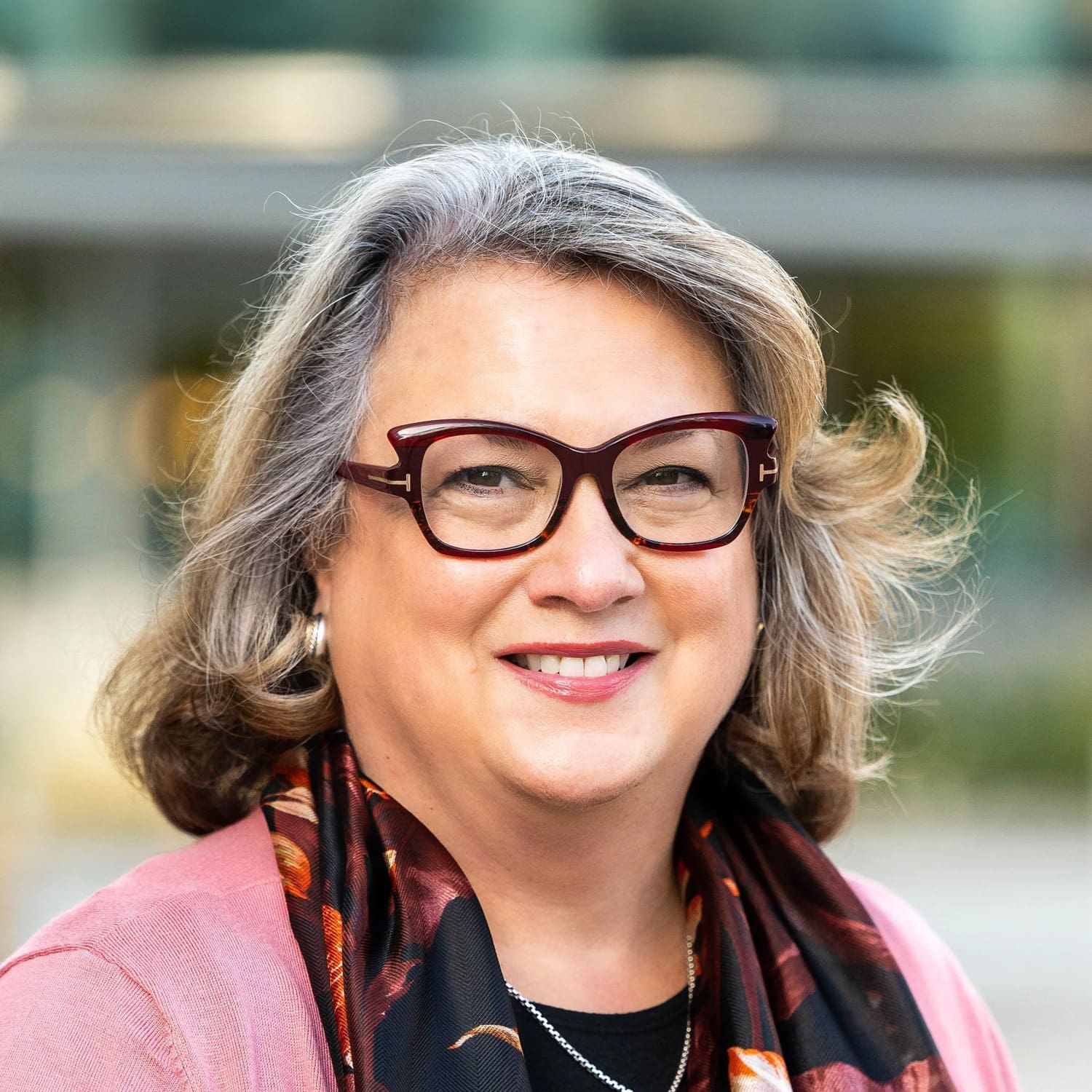 A smiling woman with shoulder-length gray hair, wearing glasses, a black and orange scarf, and a pink sweater, stands outdoors with a blurred background.