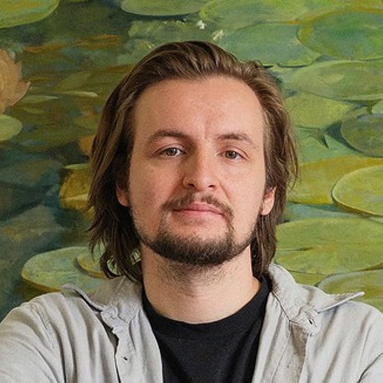 A person with medium-length brown hair and a beard stands in front of a green lily pad painting, wearing a light gray shirt over a black t-shirt, looking at the camera with a neutral expression.