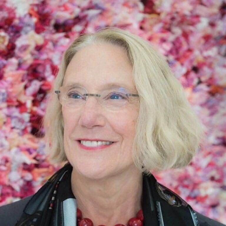 A smiling older woman with blonde hair and glasses stands in front of a vibrant, pink floral background. She wears a black scarf and a chunky red necklace.