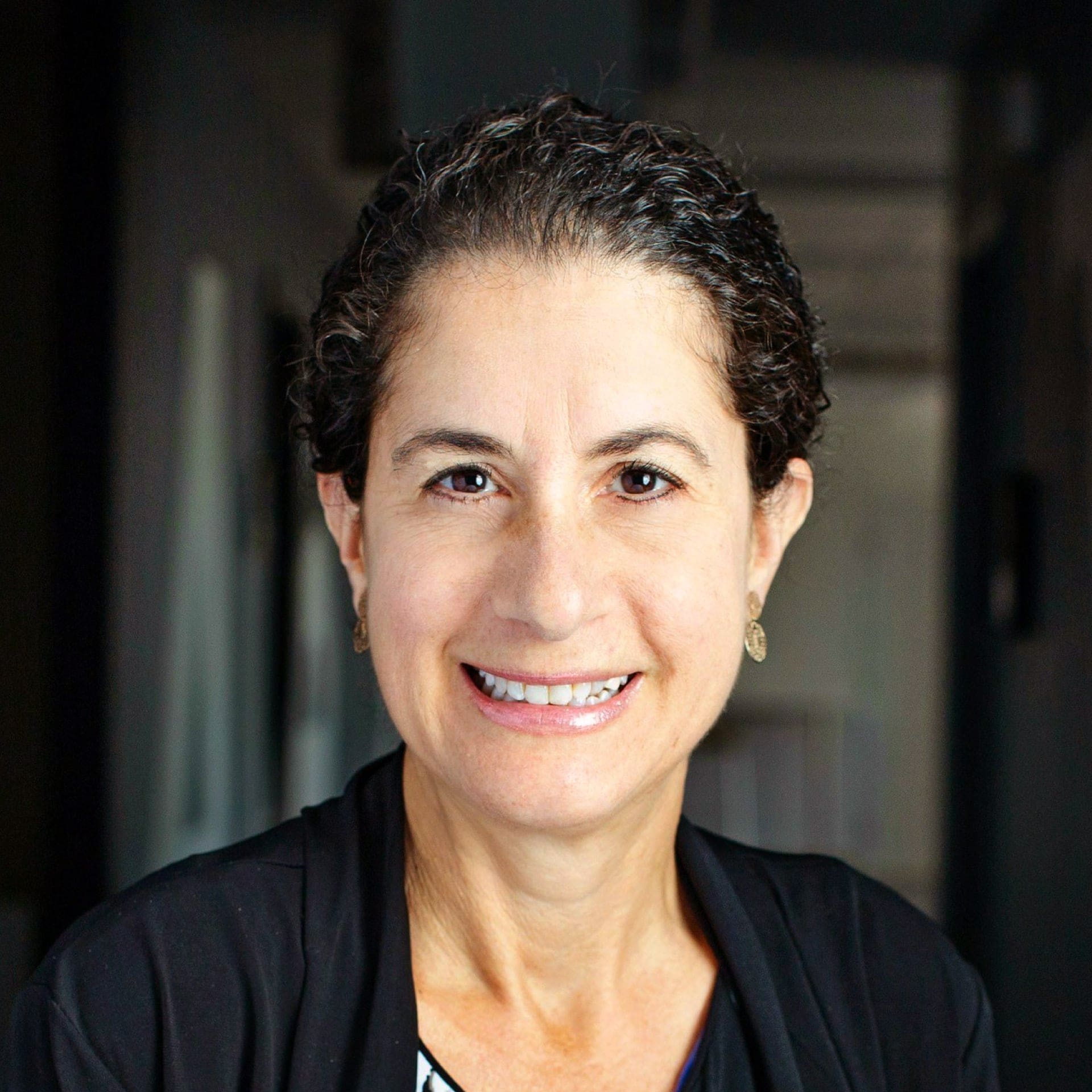 A woman with short, curly dark hair smiles at the camera, wearing a dark jacket and small earrings, with a blurred indoor background.