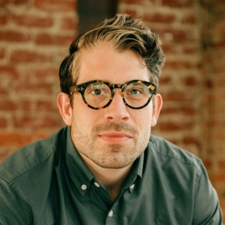 A man with short brown hair, glasses, and a trimmed beard wears a green shirt and looks at the camera. He is in front of a brick wall background.