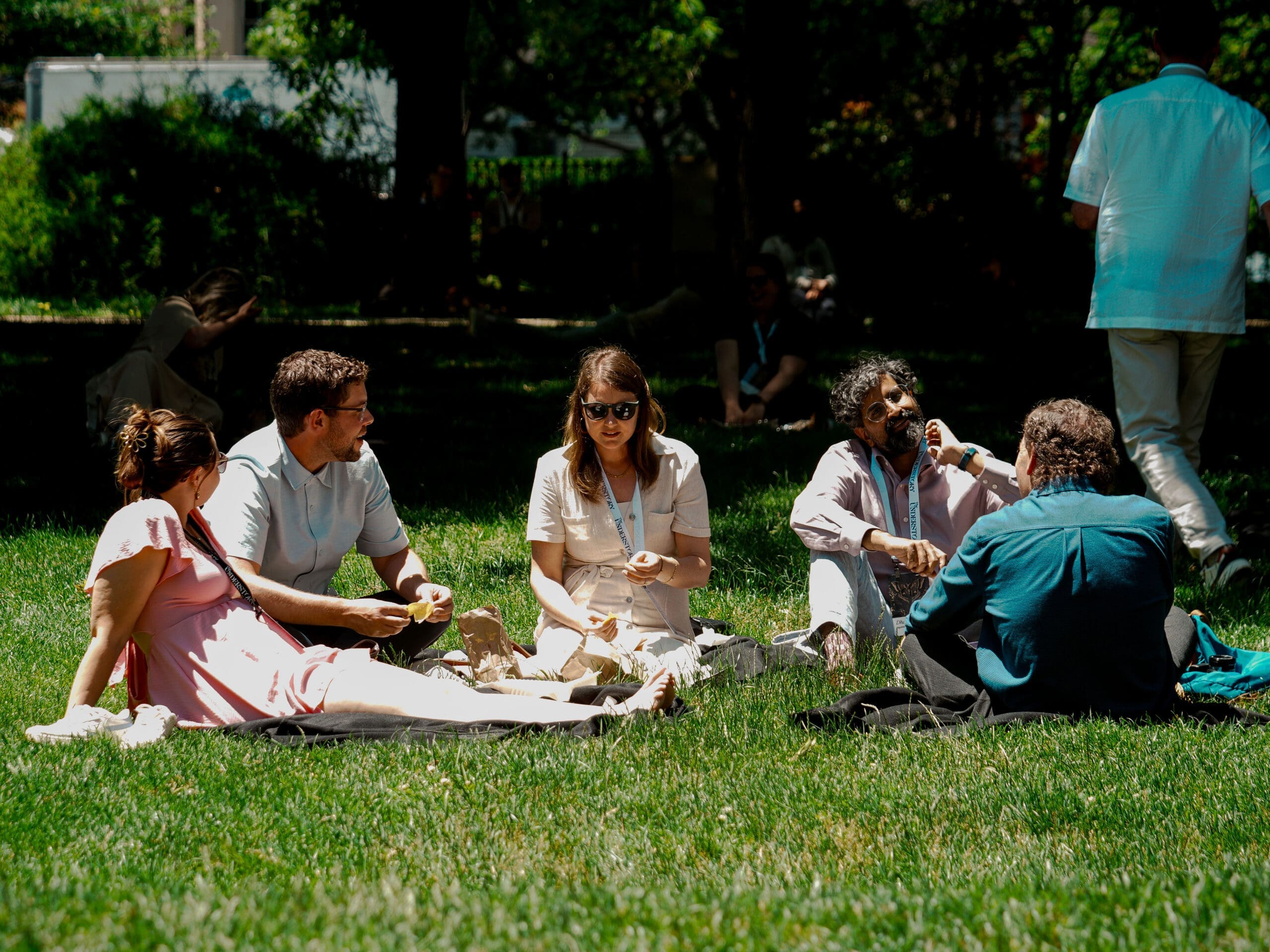 A group of people sit on the grass in a park, talking and eating together on a sunny day. Trees and other people are visible in the background.