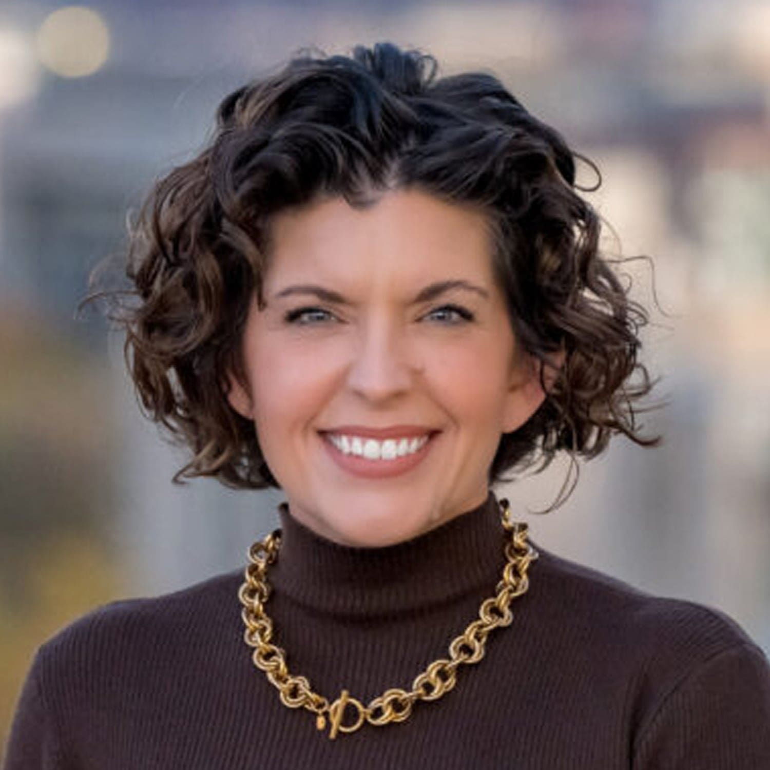 A woman with shoulder-length curly dark hair smiling, wearing a brown turtleneck and a gold chain necklace.