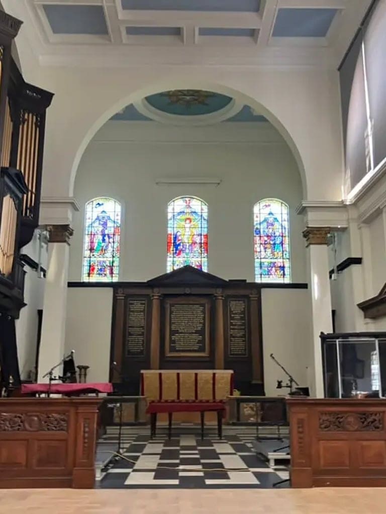 The interior of a church featuring a black and white tiled floor, three stained glass windows, and a dark wooden altar area.