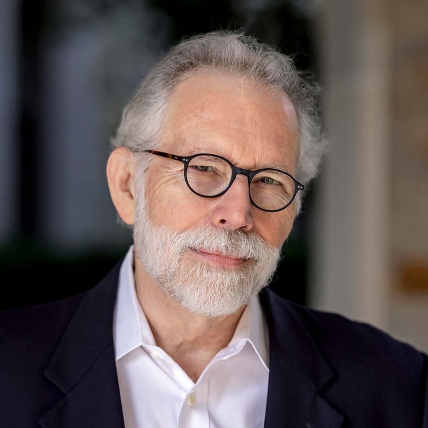 Headshot of an older man with gray hair and a beard, wearing glasses, a white collared shirt, and a dark blazer.