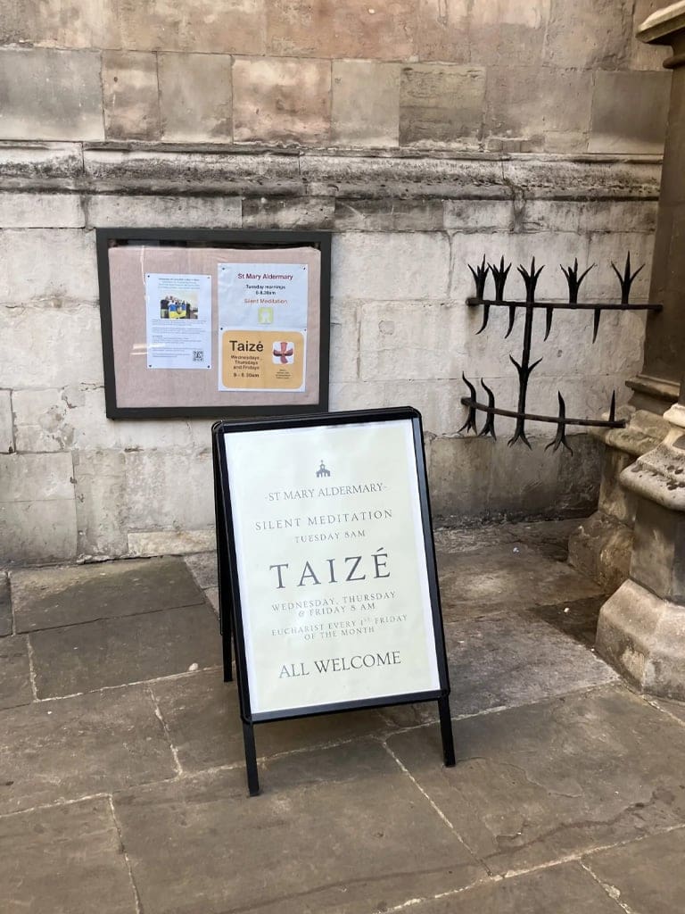 A sidewalk sign in front of a stone wall announces St. Mary Aldermarys schedule for silent meditation, Taizé services, and Eucharist, stating All Welcome.