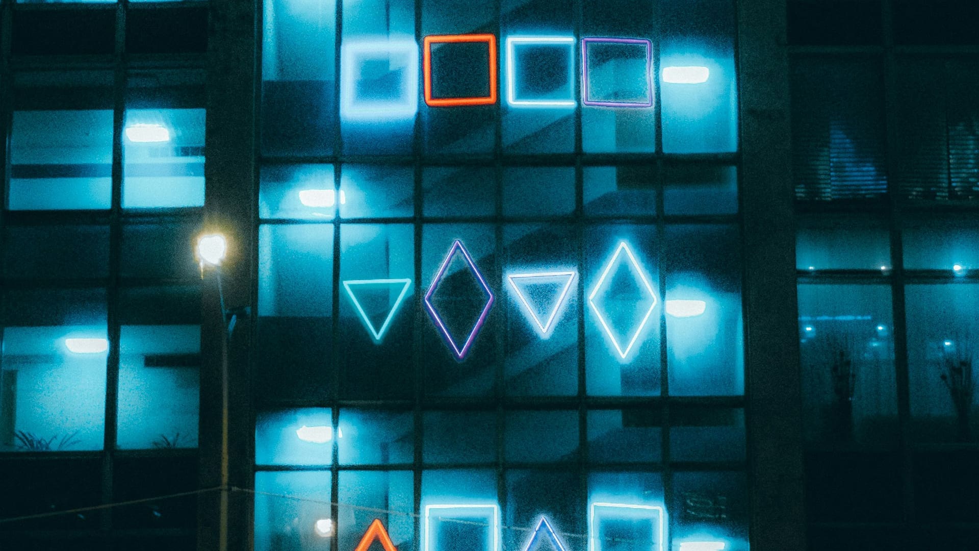 A building facade at night is lit with neon geometric shapes, including squares and diamonds, in blue and orange, against a backdrop of glass windows reflecting blue light.