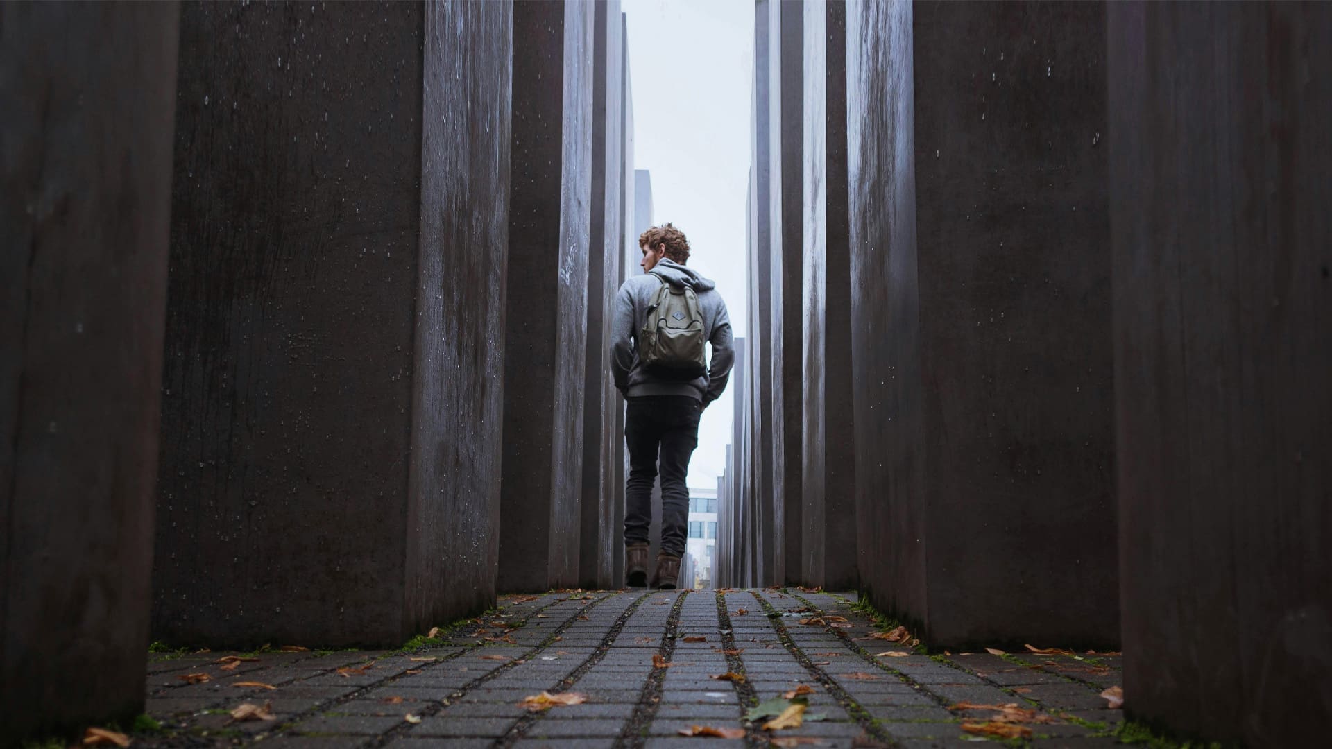A person with a backpack walks alone between tall, gray concrete slabs on a narrow pathway, with scattered leaves on the ground and diffused daylight above.