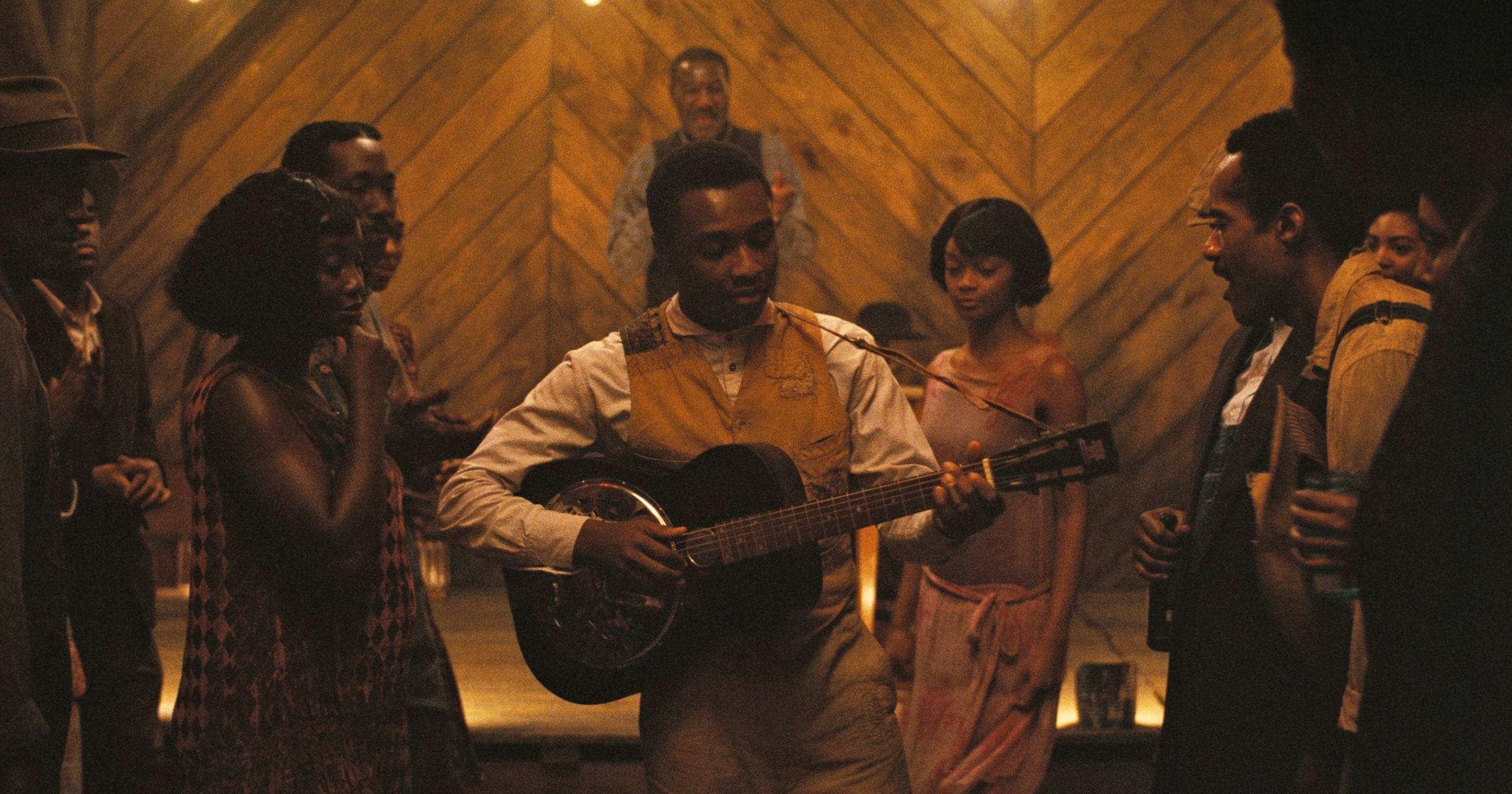 A man in vintage clothing plays an acoustic guitar and sings to a group of people gathered around him in a warmly lit, rustic room with wooden walls.
