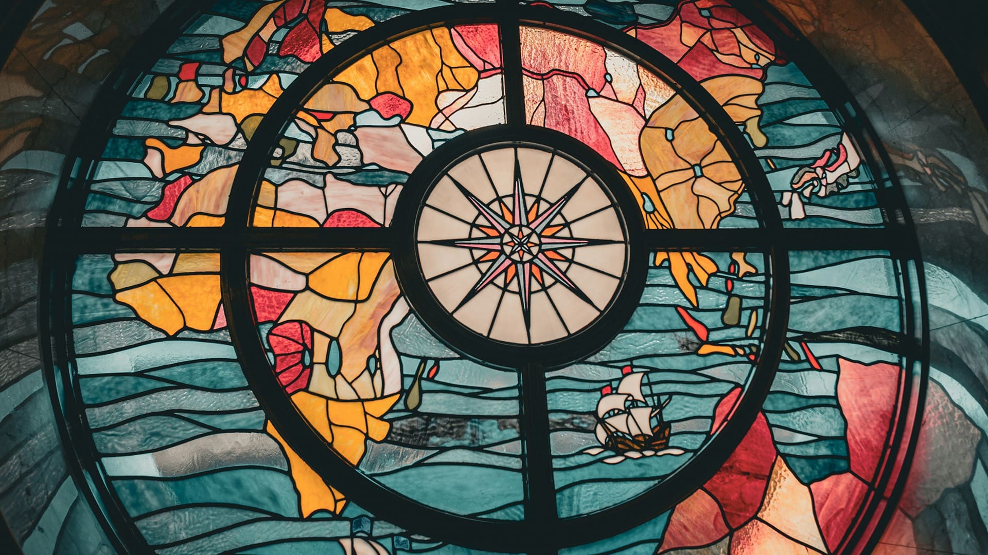 A decorative stained glass window depicts a world map with vibrant shades of blue, red, and yellow, featuring a central compass rose and a small sailing ship on the ocean.