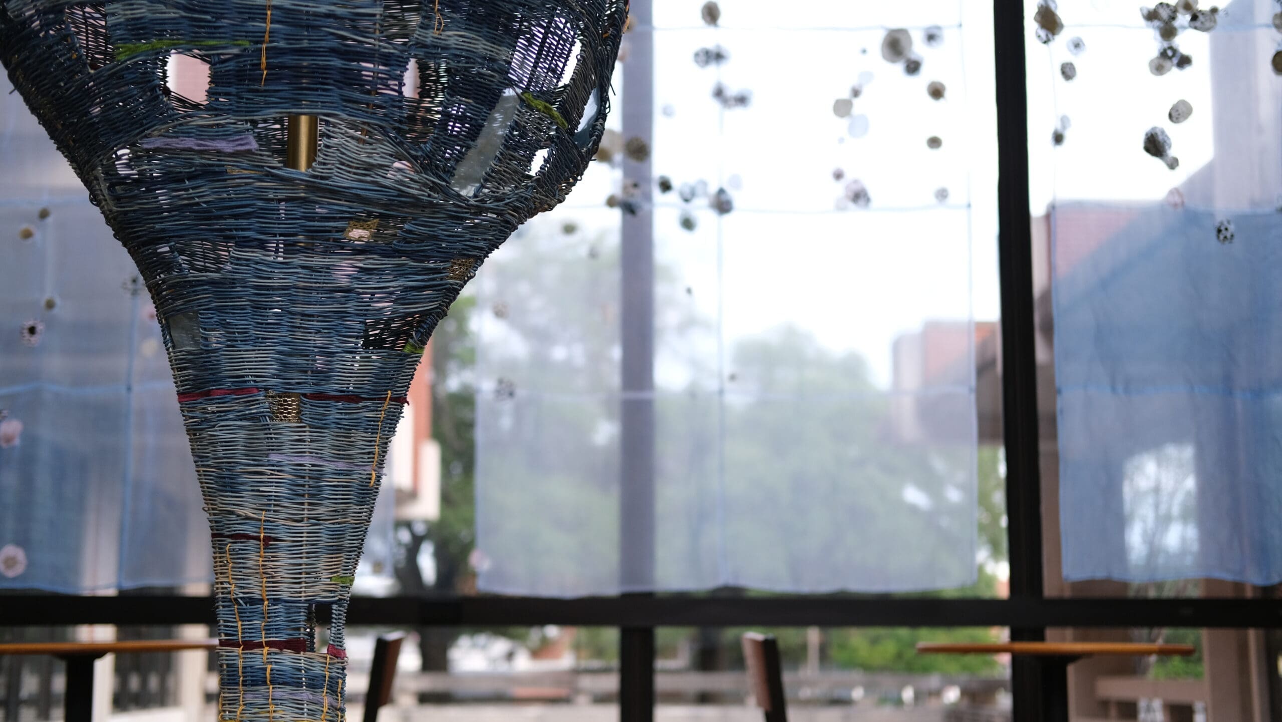 A large, intricate woven blue sculpture hangs indoors in front of tall windows with sheer blue curtains. Trees and buildings are visible outside through the glass.