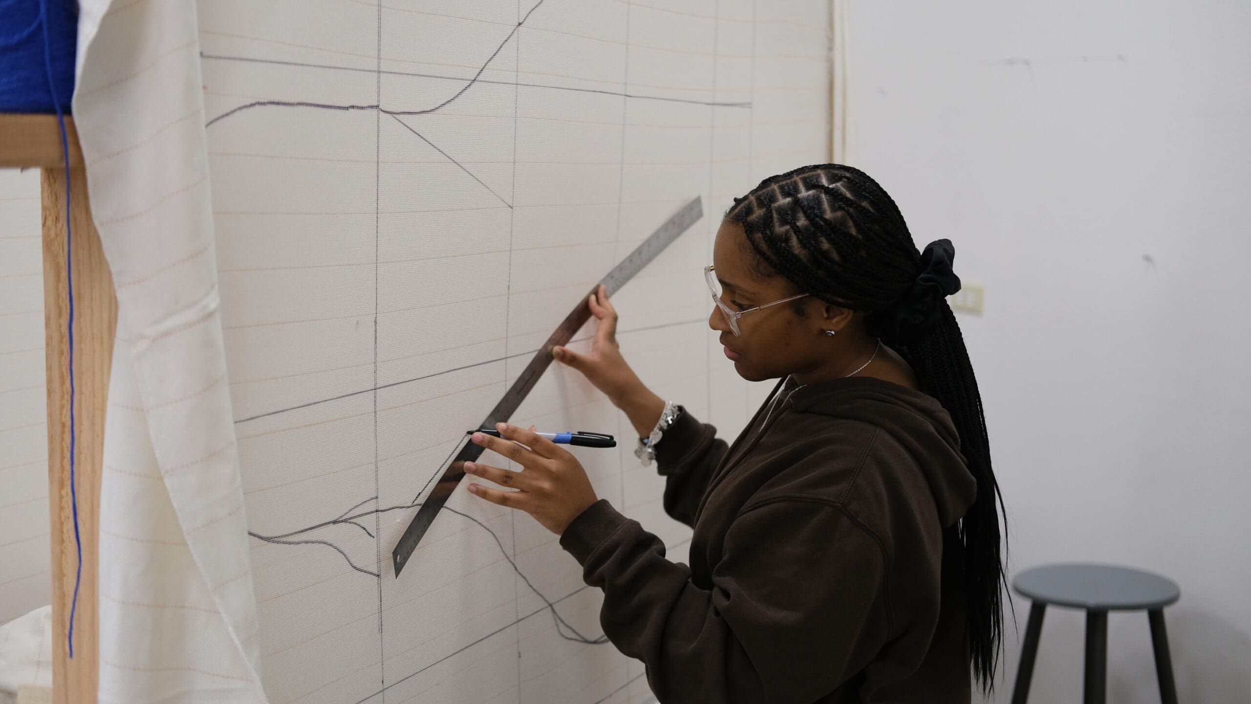 A woman wearing glasses and a brown hoodie uses a ruler and marker to draw precise lines on a large, gridded canvas mounted on a wall. A stool and some fabric are visible in the background.