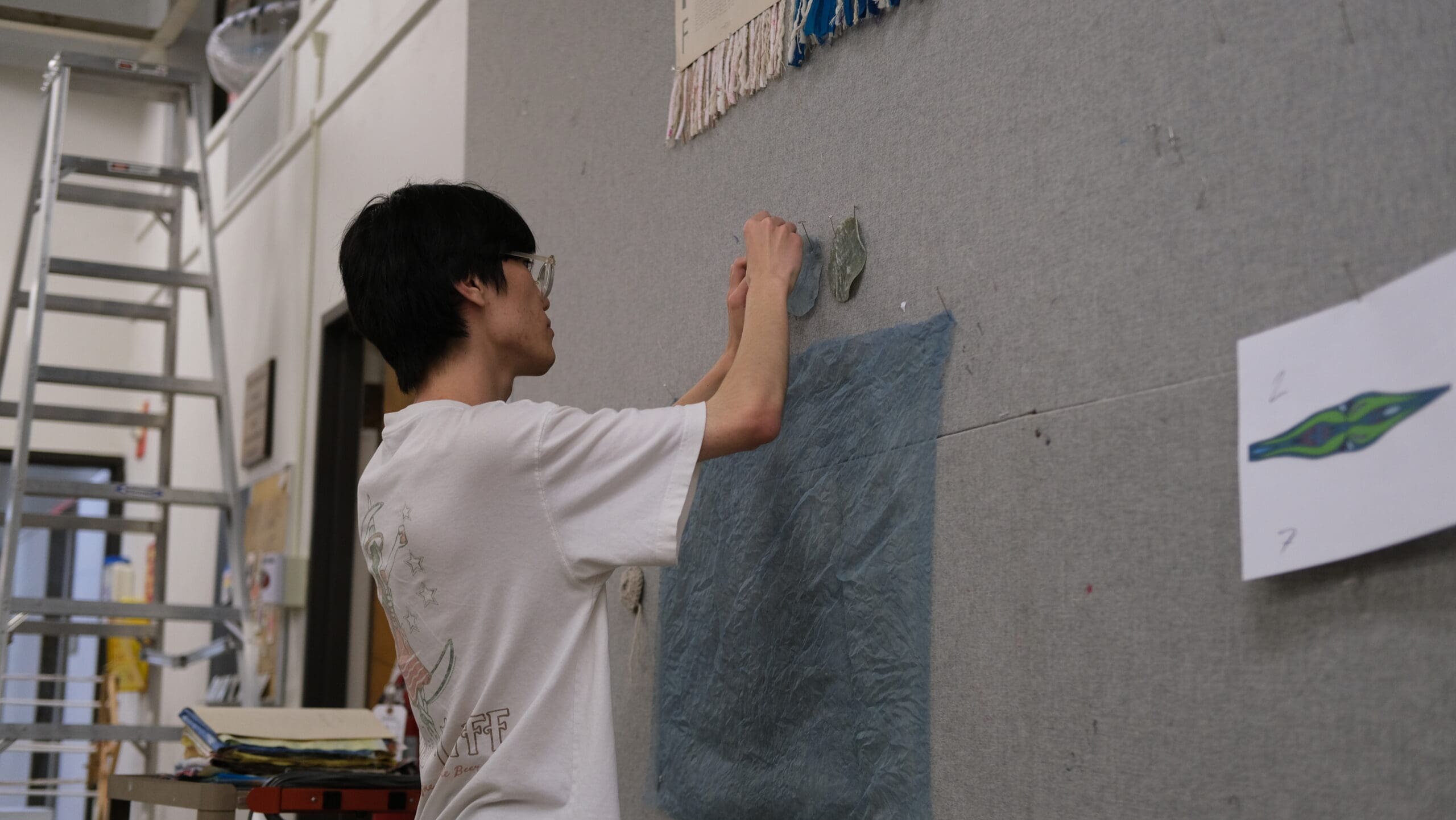 A person wearing glasses and a white shirt attaches something to a wall covered with fabric and drawings. A ladder and art supplies are visible in the background.
