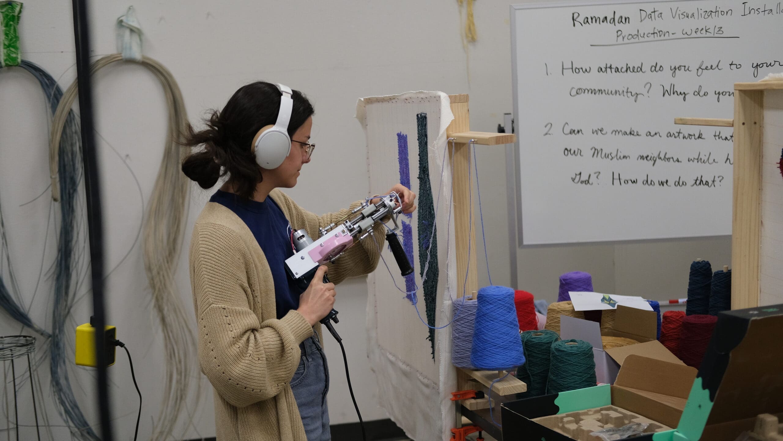 A person wearing headphones uses a tufting gun on a vertical canvas, surrounded by colorful yarn spools. In the background, a whiteboard displays project notes and questions about a Ramadan data visualization installation.