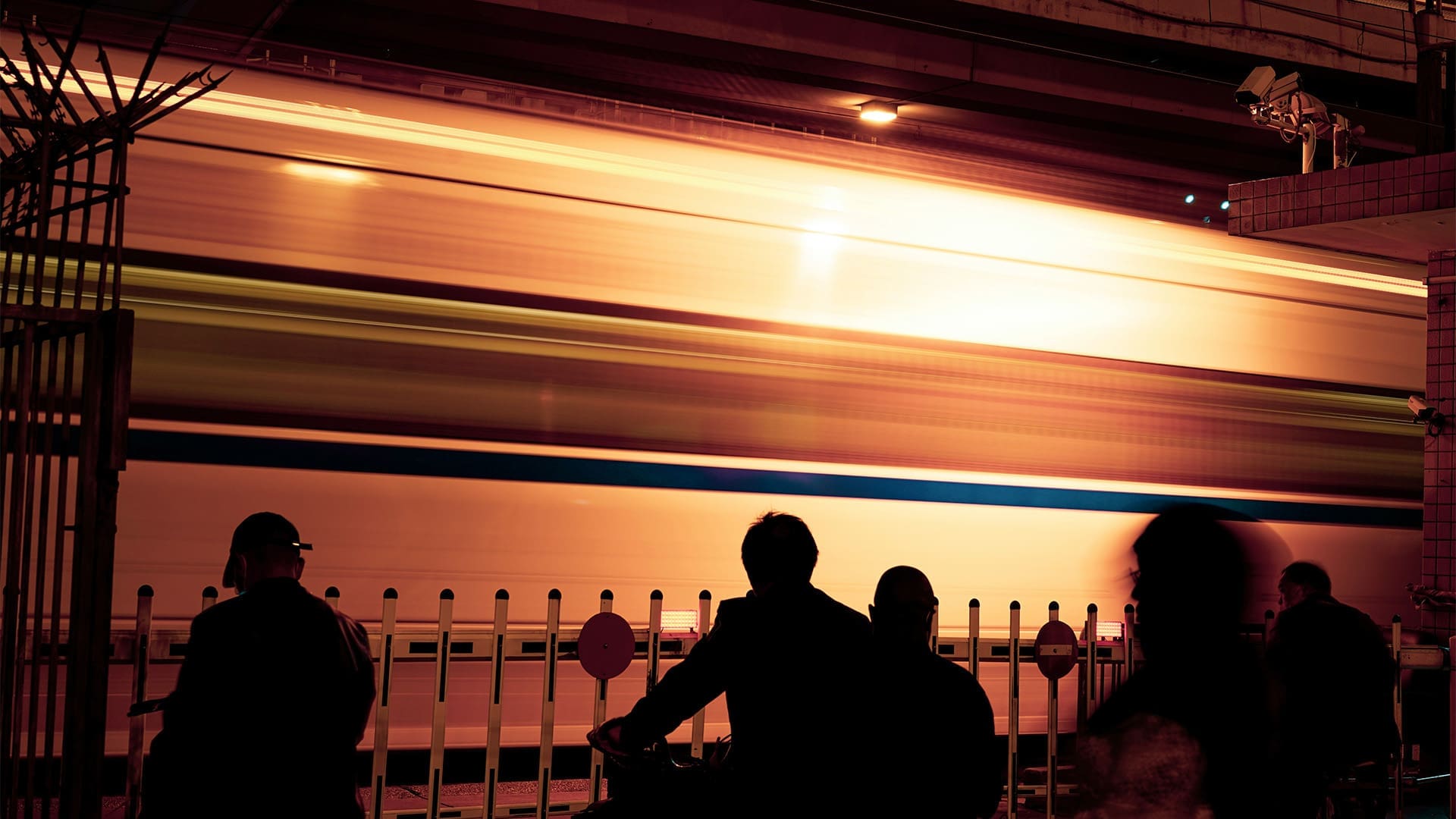 Silhouettes of people stand near a fence at night as a brightly lit train speeds past, creating a motion blur effect. The scene is illuminated by warm, orange streetlights.