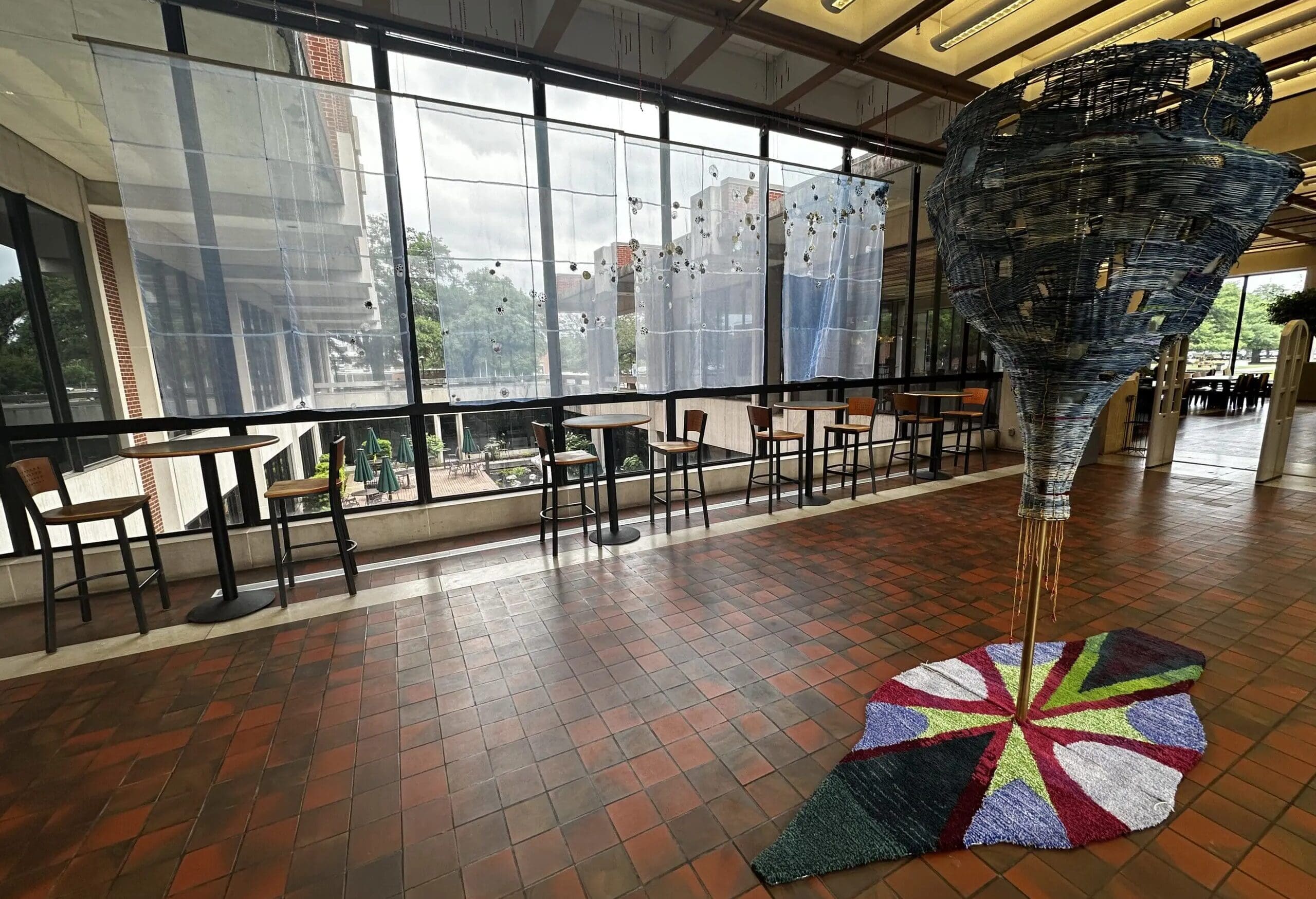 A spacious indoor area with tiled floors, high windows, tall chairs and tables, and a large, cone-shaped woven sculpture with a colorful circular base on the floor. Trees and buildings are visible outside.