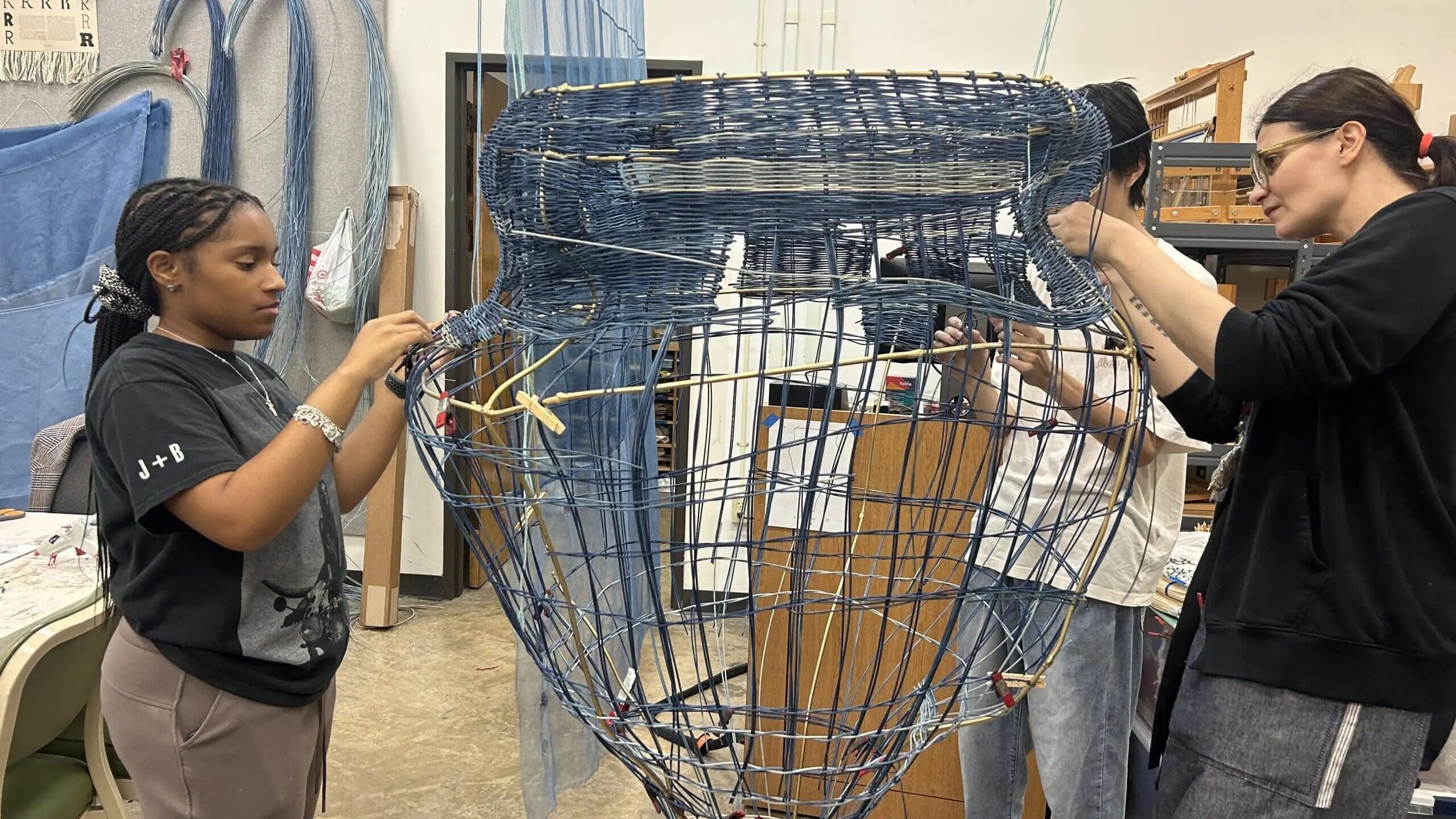 Three people work together weaving and assembling a large, blue wire sculpture shaped like a vessel in an art studio filled with materials and tools.