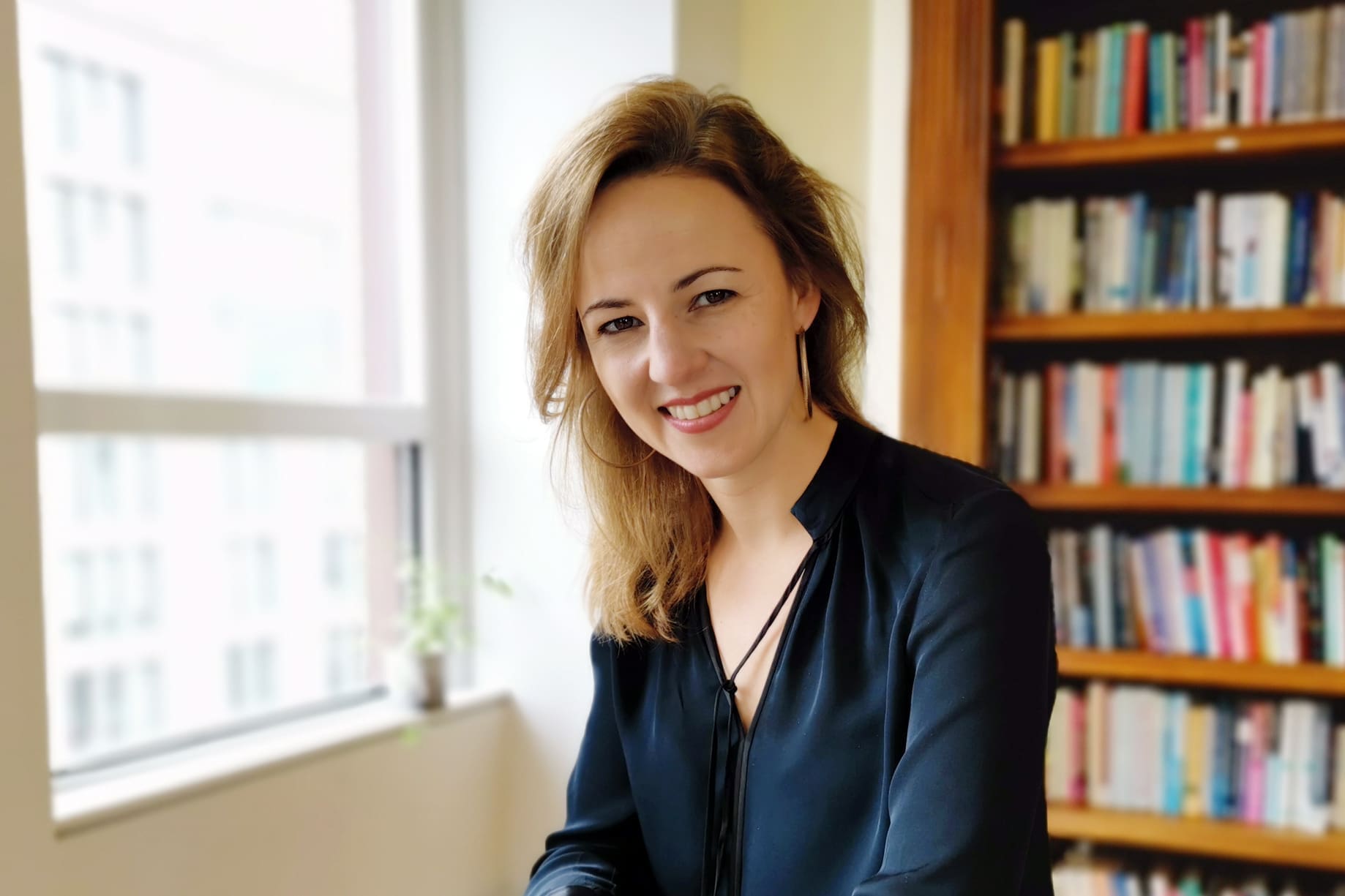 A woman with light brown hair, wearing a dark blouse, smiles while sitting in a bright room with a window and a bookshelf filled with books in the background.