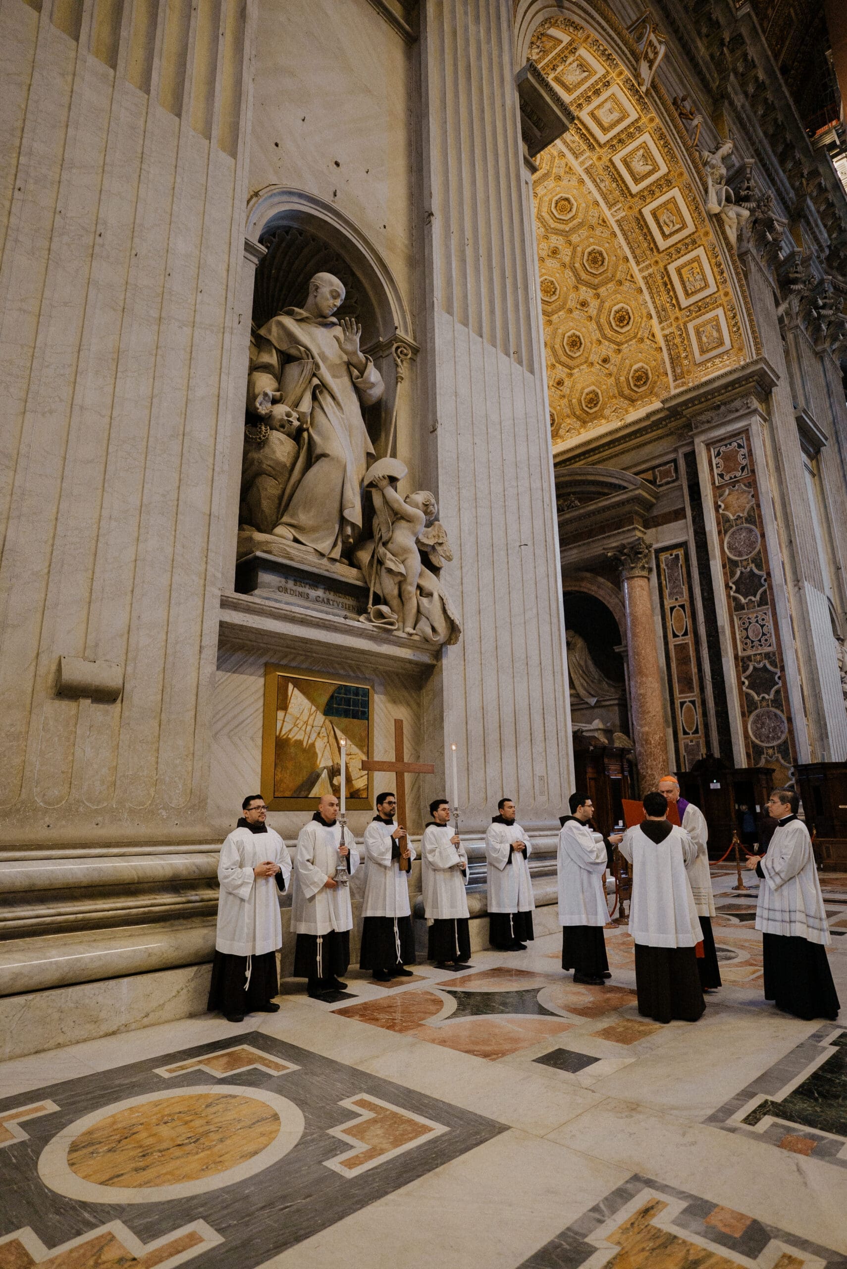 A group of clergy in white robes stands in a semicircle inside a grand, ornate church with tall columns, marble floors, and religious statues.