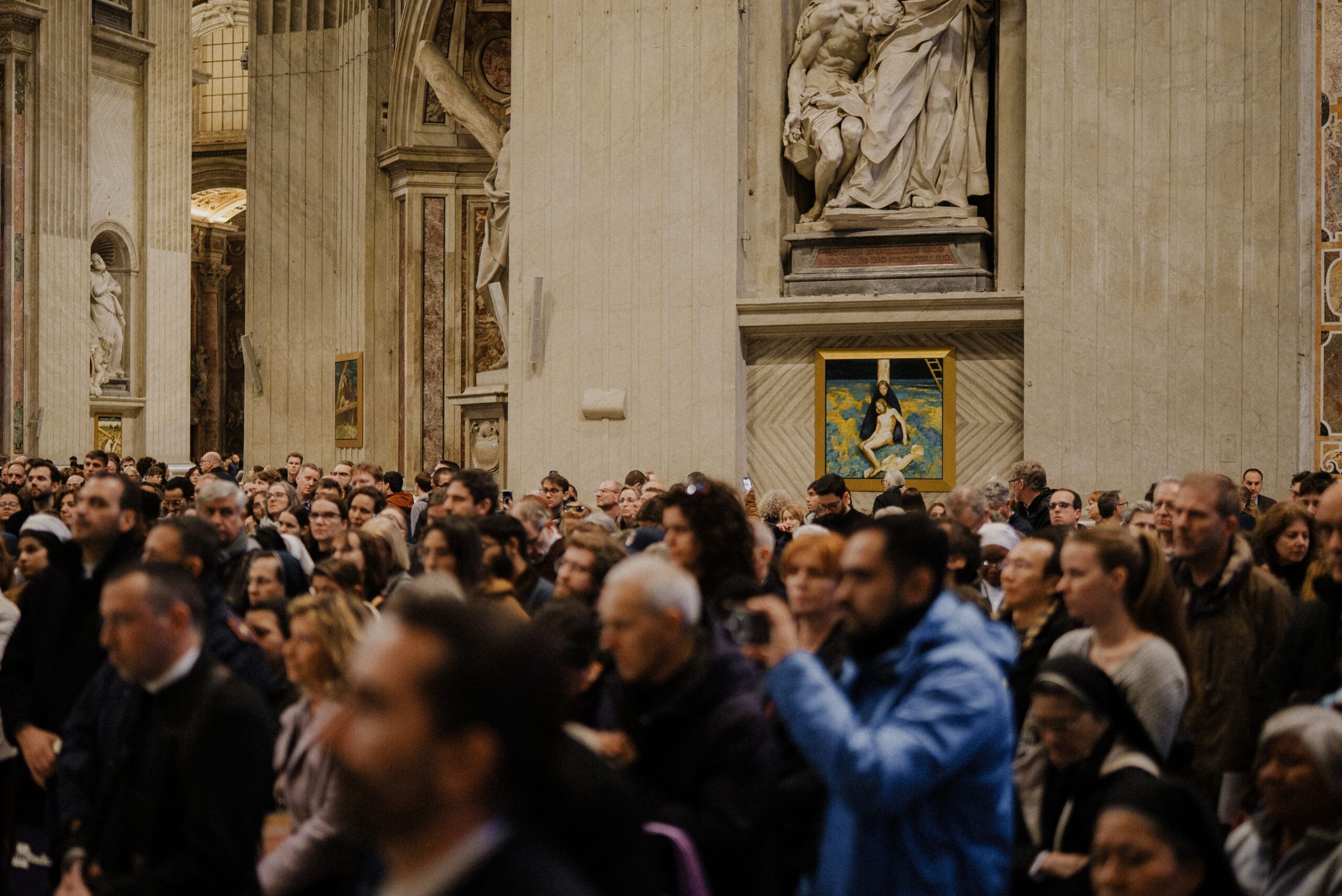 A large crowd of people gathered inside a grand, ornate church with tall columns, religious statues, and paintings on the walls. The atmosphere appears solemn and attentive.