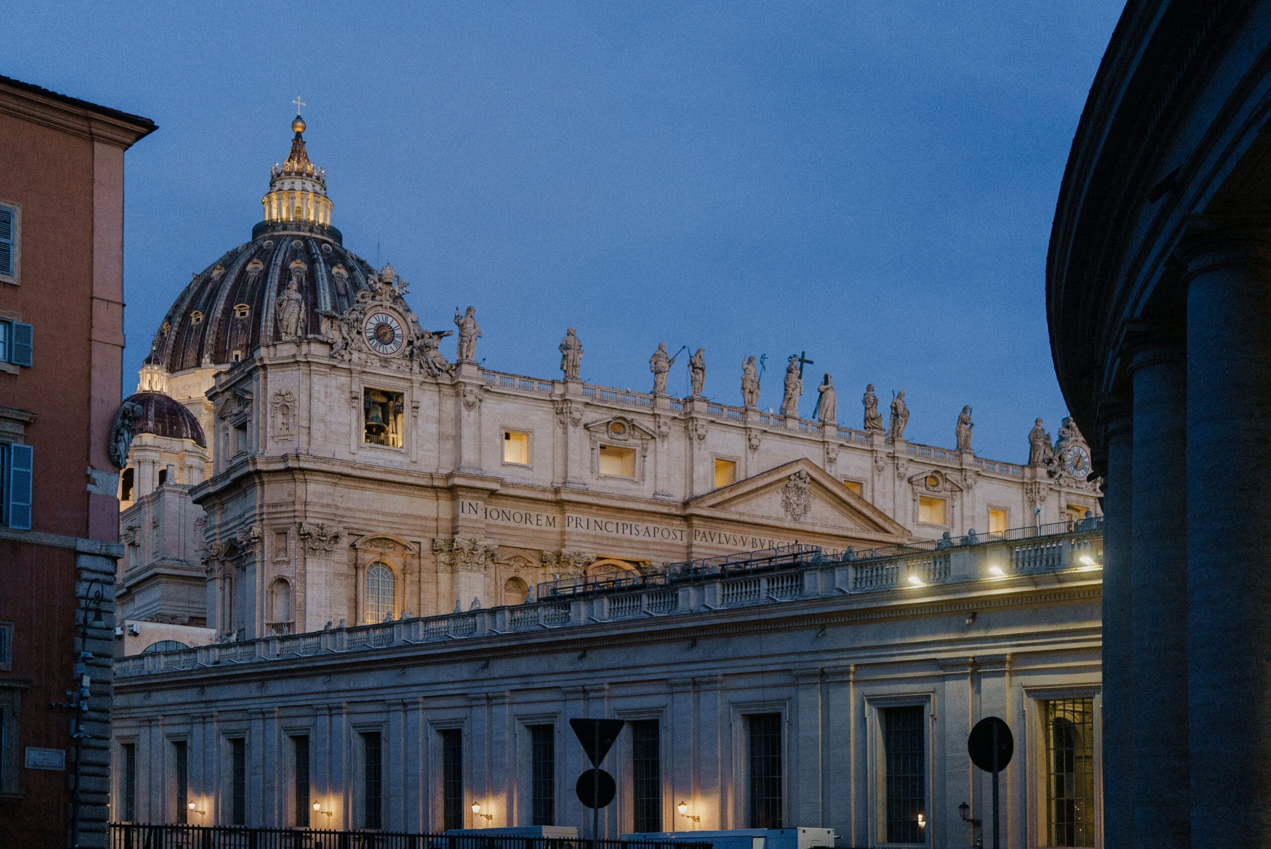 St. Peter’s Basilica in Vatican City at dusk, illuminated by lights, with statues along the roofline and a large dome visible against a blue evening sky.