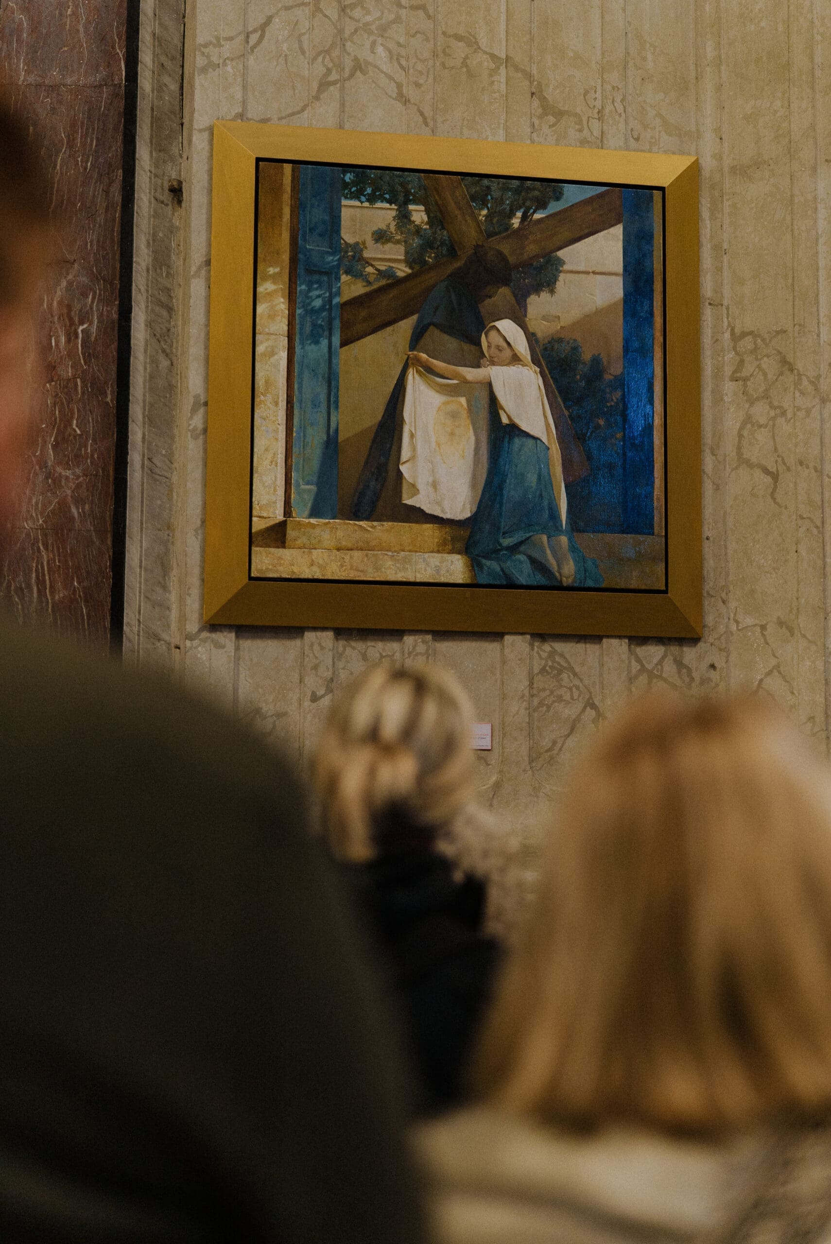 A framed painting of a religious scene featuring two figures near a cross hangs on a marble wall, with several people viewing it from below.