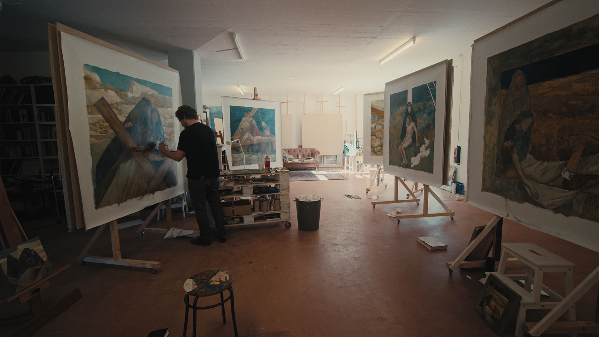 An artist stands in a spacious, well-lit studio working on a large painting. Three other large artworks are displayed on easels around the room, and art supplies are scattered on the floor and nearby tables.