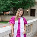 A young woman in a magenta dress and Texas graduation stole smiles while standing by a stone railing, with trees and a building in the background.