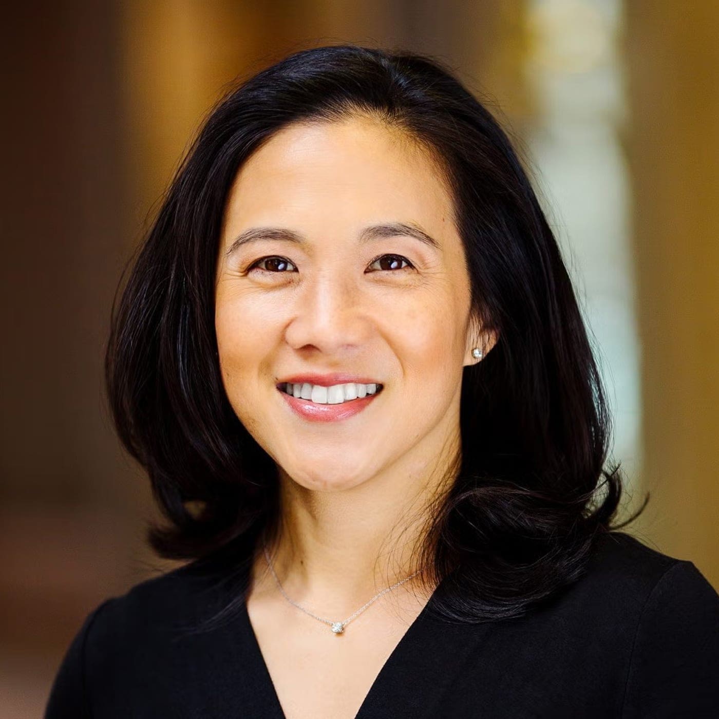 A woman with shoulder-length black hair is smiling at the camera. She is wearing a black top and a delicate necklace. The background is softly blurred in warm tones.